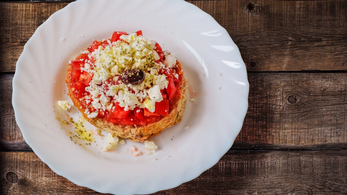 A close-up of a classic Greek Dakos salad with feta, tomato, and olive on a wooden table.